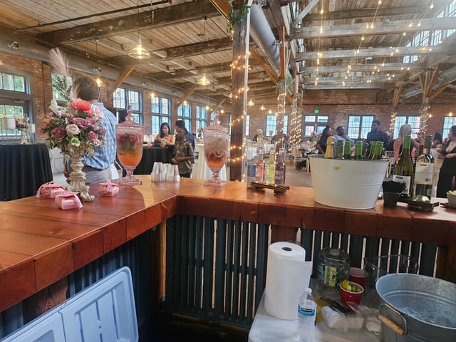 An L-shaped wood bar in a rustic mill venue with a reception under string lights